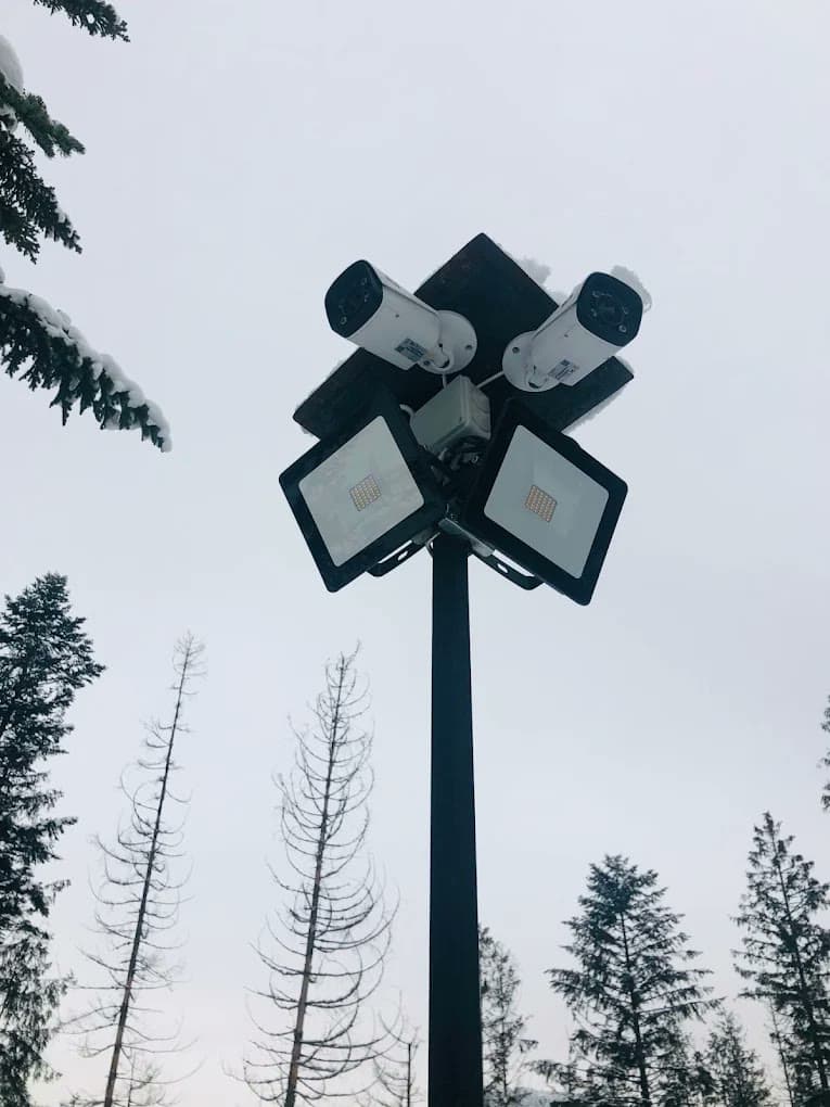 Dwie kamery IP i dwa naświetlacze LED na słupie nad parkingiem, zimą, wśród świerków